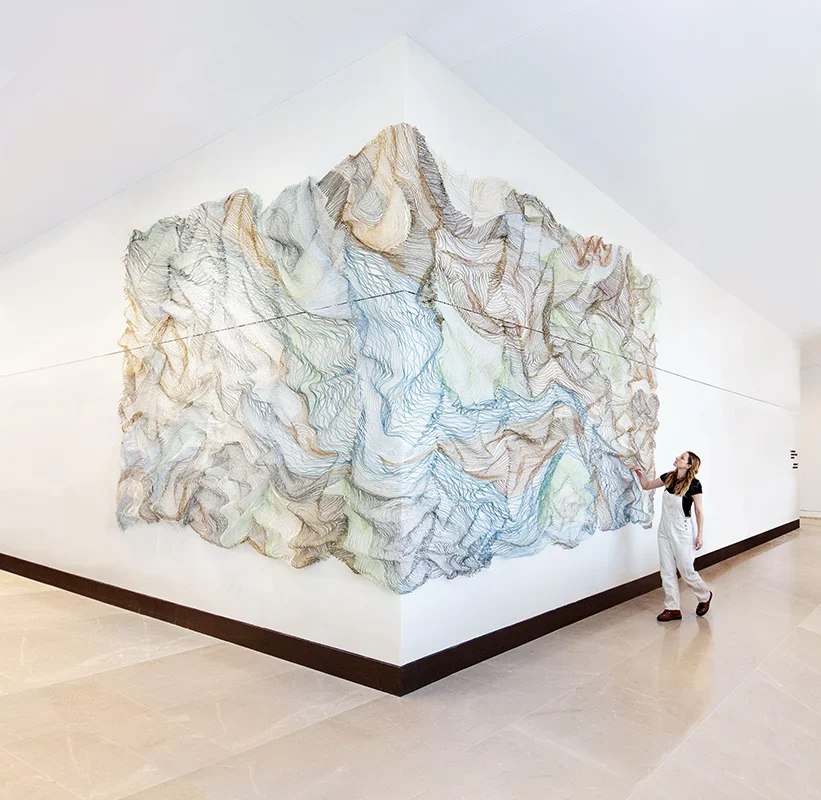 A woman closely examines an intricately woven large-scale wall sculpture in the lobby of an office.