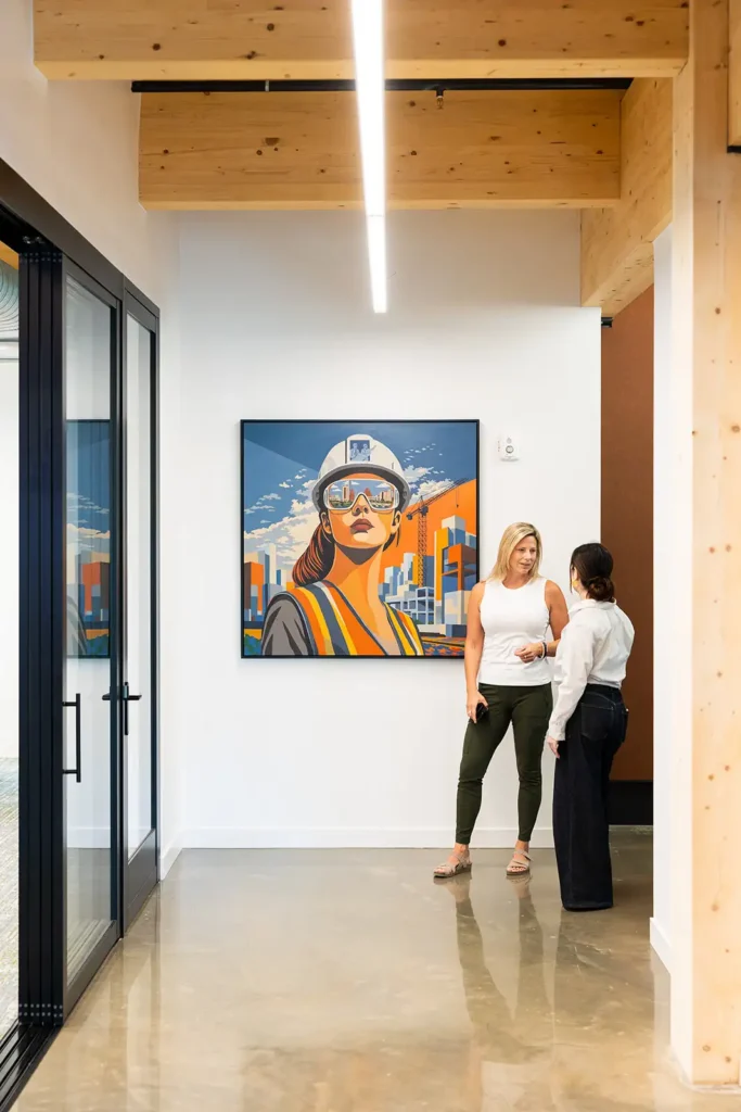 Two women converse in a hallway in front of a painting of a female construction worker.