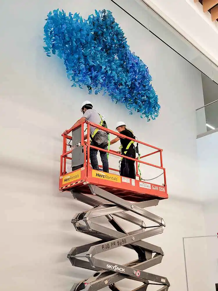 A man and a woman wearing hard hats stand on a scissor lift below a large blue textural art installation on a white wall.